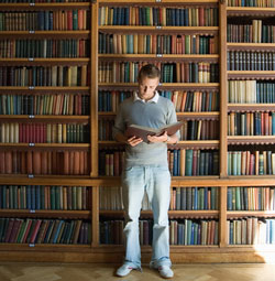 Photo of a man reading a book, standing in front of a bookshelf loaded with books.